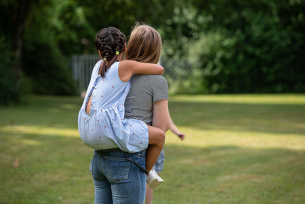 Eine junge Frau trägt ein kleines Mädchen in einem hellblauen Kleid huckepack.