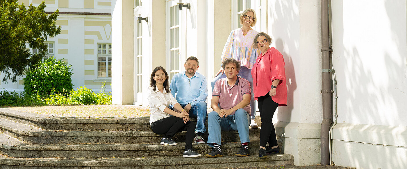 Fünf Personen in legerer Kleidung sitzen und stehen auf Treppenstufen vor einem weißen Gebäude.
