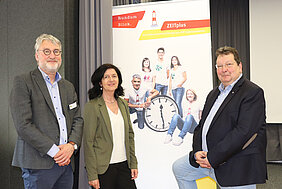 Zwei Männer und eine Frau stehen vor einem Banner, auf dem das Logo des Lebensarbeitszeitmodells ZEITplus abgebildet ist. 