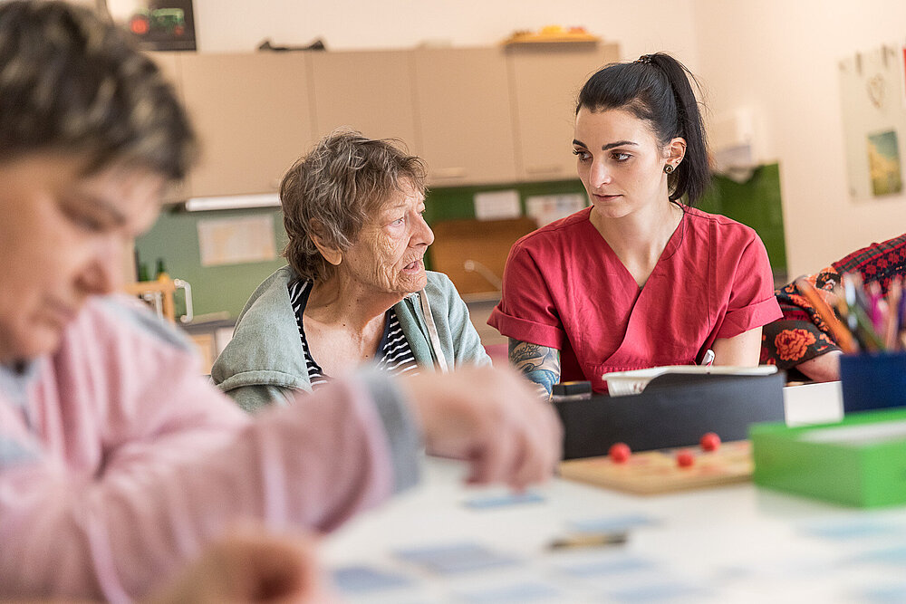 Eine junge Frau in roter Dienstkleidung sitzt mit zwei Patientinnen an einem Tisch, auf dem sich Mal- und Bastelutensilien befinden.
