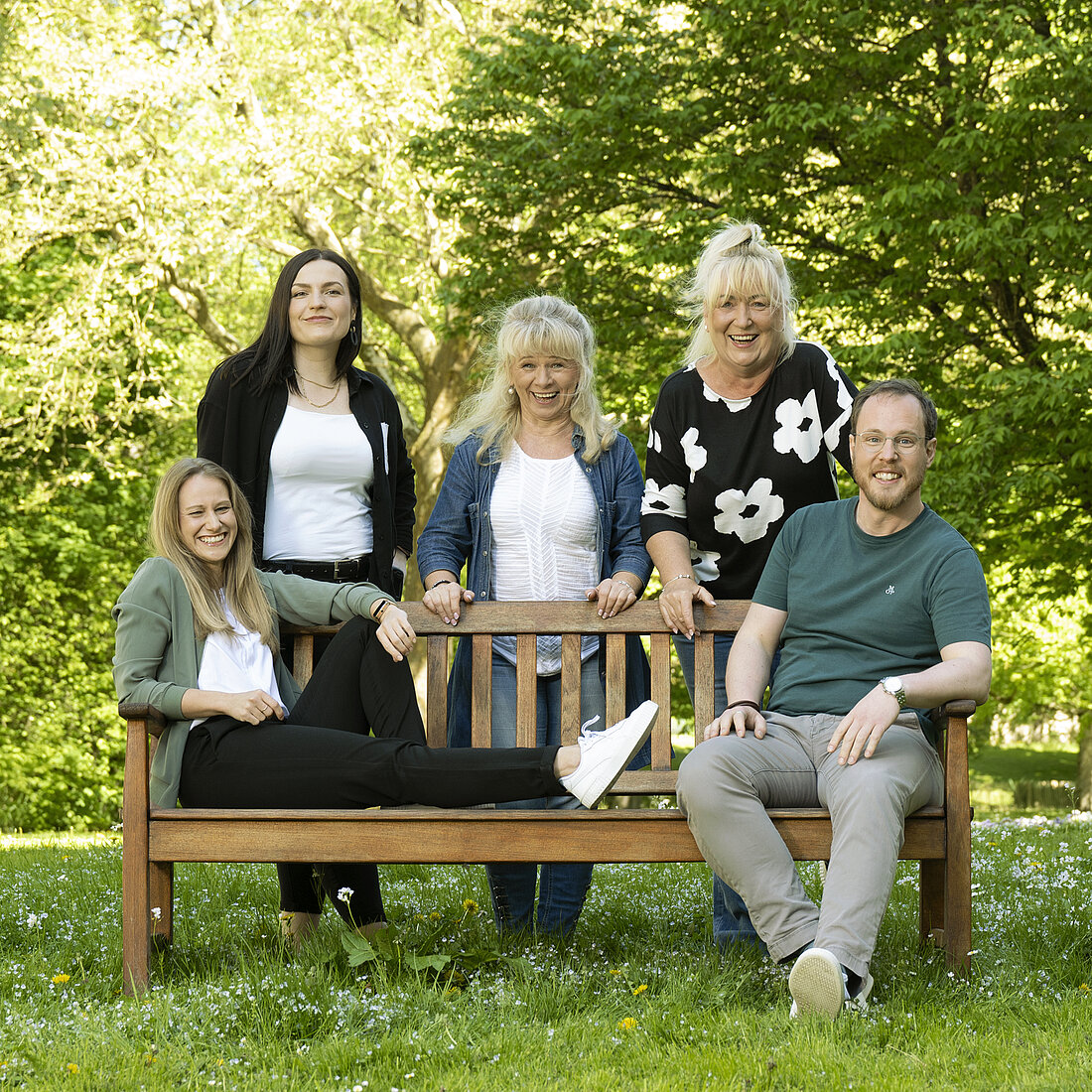 Fünf Personen in einem Park, zwei sitzen auf einer Holzbank, die anderen stehen oder sitzen daneben, umgeben von Bäumen und Gras.