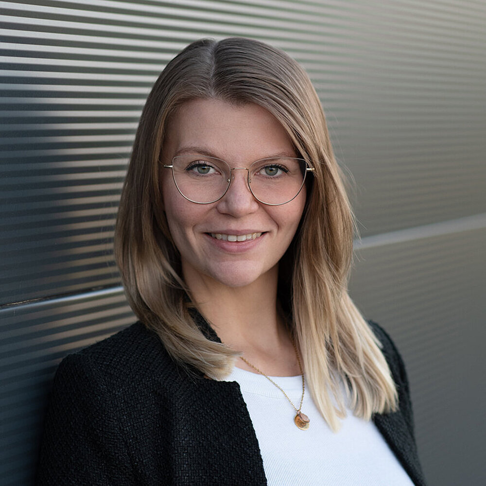 Eine Frau mit blonden langen Haaren, Brille und weißer Bluse schaut lächelnd in die Kamera.