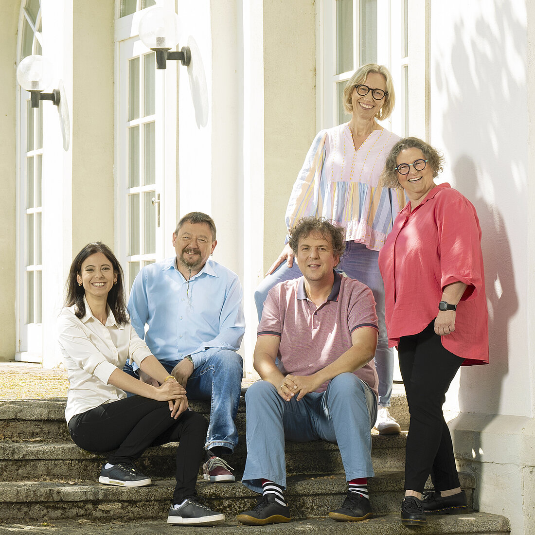 Fünf Personen in legerer Kleidung sitzen und stehen auf Treppenstufen vor einem weißen Gebäude.