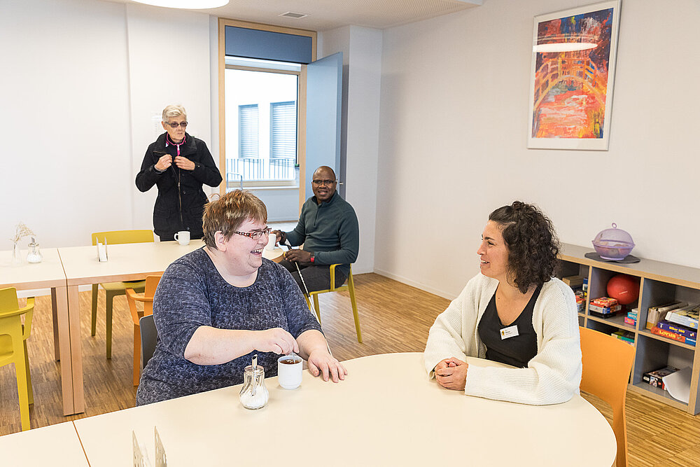 Eine Patientin und eine Mitarbeiterin der Pflege sitzen sich lachend an einem Tisch in einem Aufenthaltsraum schräg gegenüber. Die Patientin rührt in einer Kaffeetasse, im Hintergrund sind zwei weitere Personen zu sehen.