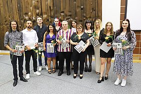 Eine Gruppe junger Menschen steht aufgereiht nebeneinander, alle halten eine Rose und eine Urkunde in den Händen. 