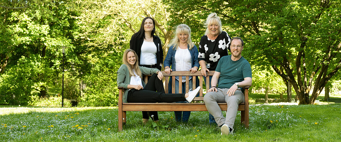 Fünf Personen in einem Park, zwei sitzen auf einer Holzbank, die anderen stehen oder sitzen daneben, umgeben von Bäumen und Gras.