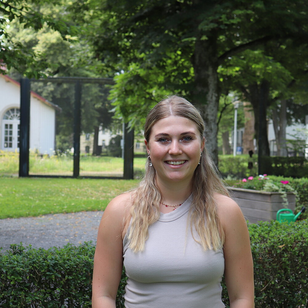 Eine junge Frau mit langen blonden Haaren steht im Freien und lacht in die Kamera, hinter sich Wiese, Bäume und viel grün.
