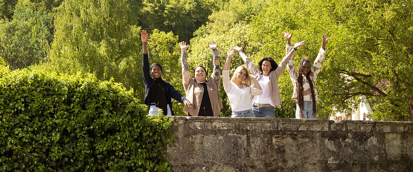 Fünf Personen stehen hinter einer Steinmauer in einem grünen Park und heben die Arme jubelnd in die Luft.