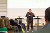Ein Mann mit Brille hält eine Präsentation auf einer Tagung. Er spricht vor einem Publikum und verwendet eine Präsentationsfolie.