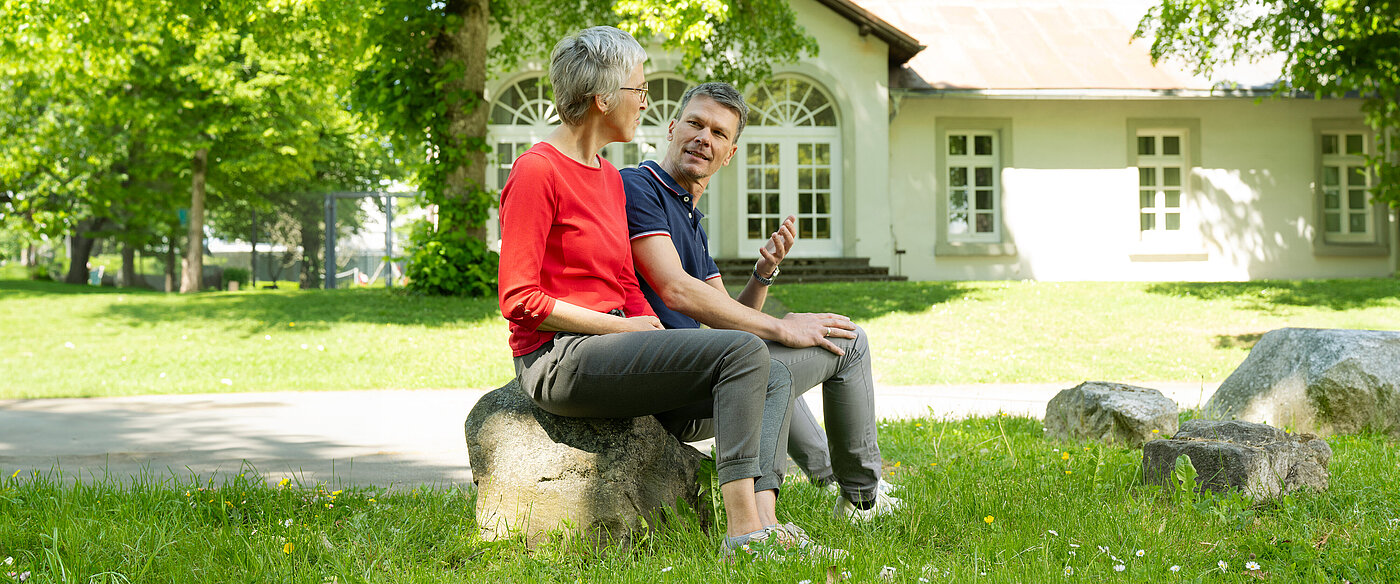 Zwei Personen sitzen auf einem Stein im Garten vor einem weißen Haus mit großen Fenstern, unter einem Baum im Schatten.