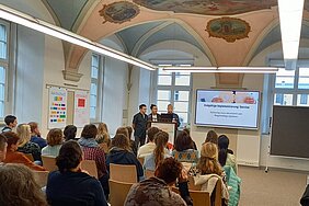 In einem historischen Klostergebäude referieren drei junge Männer vor Publikum. Sie stehen neben einer Beamer-Leinwand.