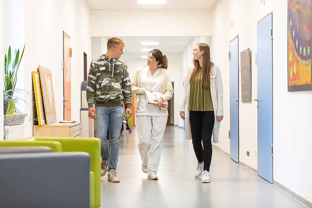 Drei junge Menschen gehen einen Gang entlang. Die Frau in der Mitte trägt Arbeitskleidung und eine Mappe, der Mann links und die Frau rechts Alltagskleidung.