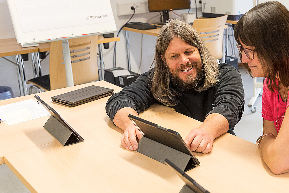 Ein Mann mit Bart erklärt zwei Frauen die Funktionsweise eines Laptops. 