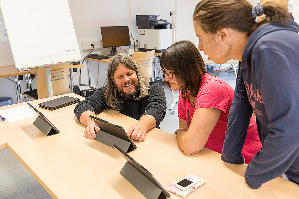 Ein Mann mit Bart erklärt zwei Frauen die Funktionsweise eines Laptops.