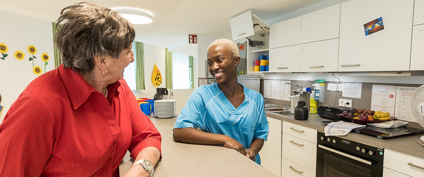 Eine junge Frau in blauer Arbeitskleidung stützt sich mit dem Arm auf einer Theke ab. Ihr gegenüber steht in ähnlicher Position eine ältere Frau mit einem roten Pullover. Die beiden lächeln sich an.