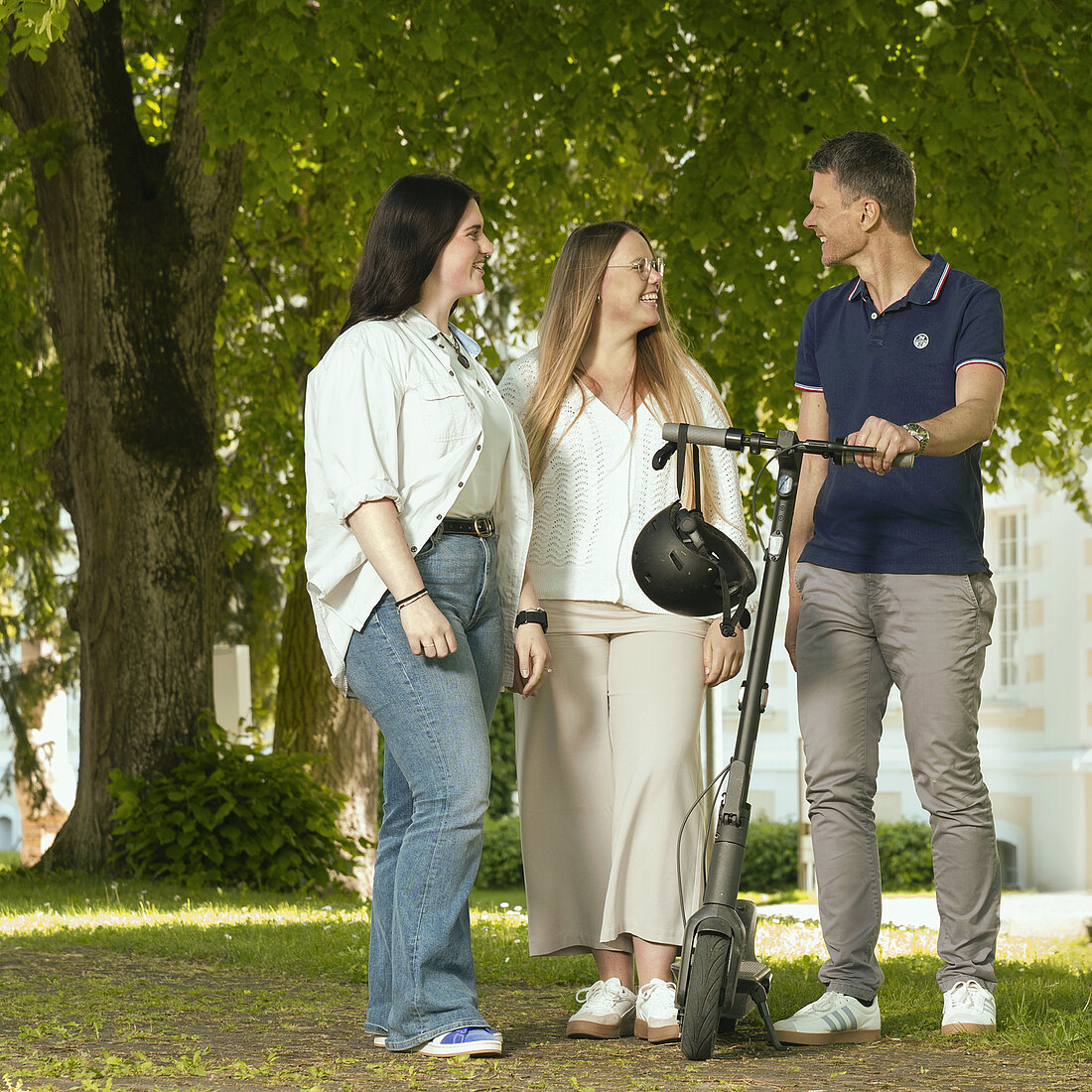 Drei Personen stehen im Park, eine hält einen E-Scooter mit Helm, alle tragen legere Kleidung.