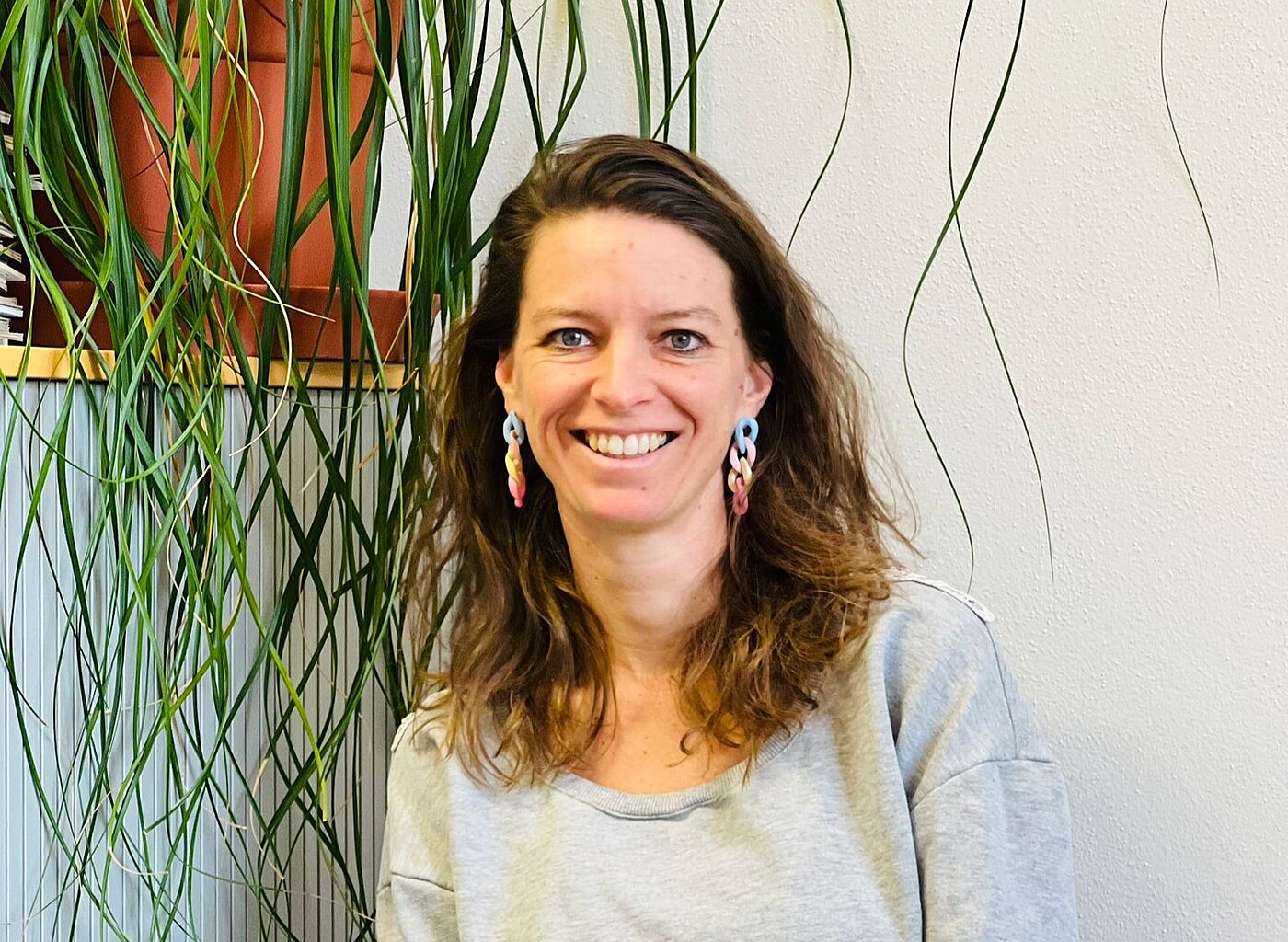 Eine Frau mit schulterlangen offenen Haaren und großen Ohrringen sitzt lächelnd vor einer Zimmerpflanze.