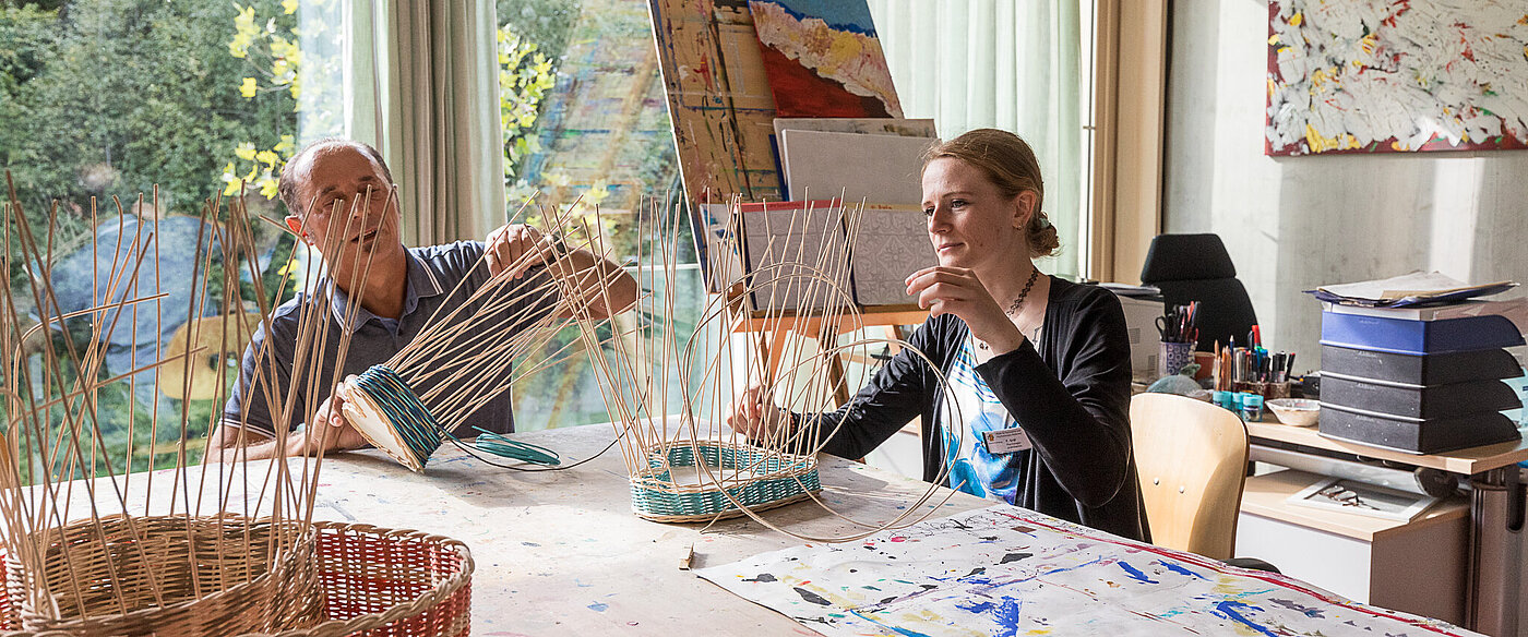 Ein Mann und eine Frau sitzen an einem großen Werktisch und flechten Körbe aus Peddingrohr. Im Vordergrund ist ein halbfertiger Korb zu sehen.