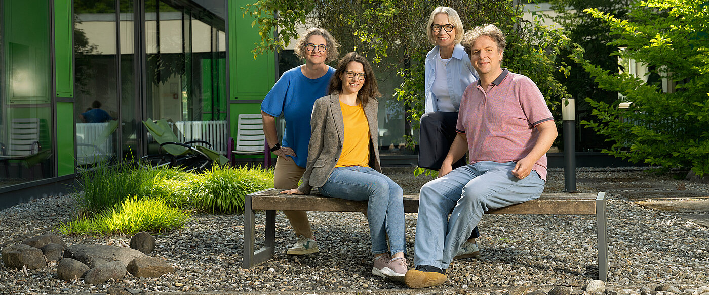 Vier Personen sitzen und stehen entspannt auf einer Holzbank im Außenbereich eines modernen Gebäudes mit grünen Fassadenelementen.