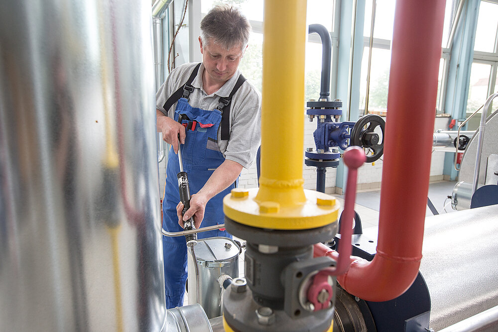 Ein Mann in blauer Latzhose hat ein Werkzeug in der Hand. Im Vordergrund sind dicke Rohre zu sehen.  
