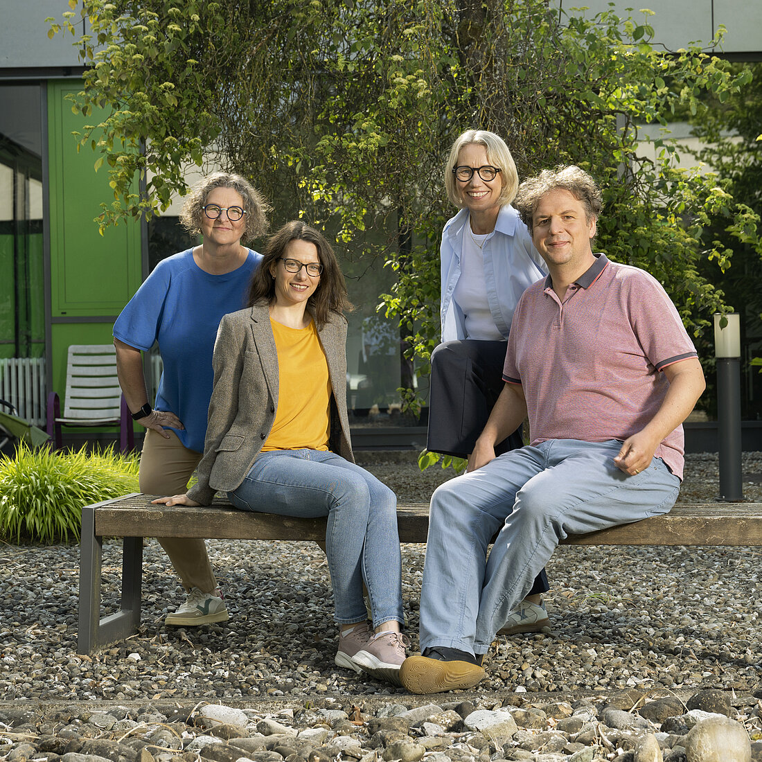 Vier Personen sitzen und stehen entspannt auf einer Holzbank im Außenbereich eines modernen Gebäudes mit grünen Fassadenelementen.