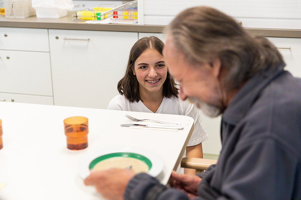 Eine junge Auszubildende aus der Pflege beugt sich zu einem Patient hinunter, der am Tisch isst. Beide schmunzeln.