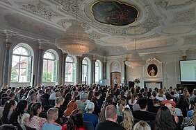 Ein vollbesetzter und bestuhlter Klostersaal, die Teilnehmenden sind von hinten und seitlich zu sehen und blicken auf eine kleine Bühne nach vorne, wo zwei Personen moderieren.