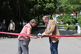 Eine Frau und ein Mann öffnen die Schleife eines roten Bandes, das quer über einen Weg gespannt ist.