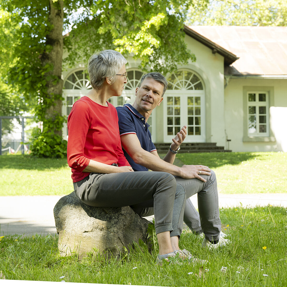 Zwei Personen sitzen auf einem Stein im Garten vor einem weißen Haus mit großen Fenstern, unter einem Baum im Schatten.